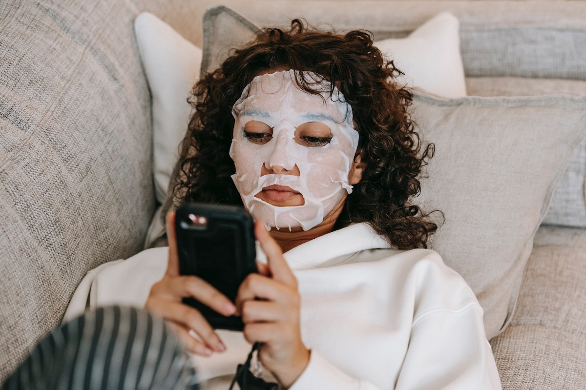 A woman browsing on a smartphone while lying on a couch in a hydrating facial mask