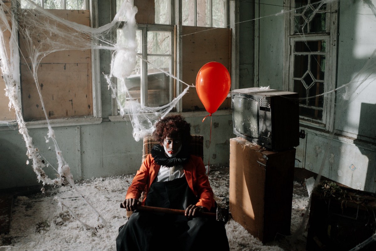 A person in clown makeup sitting with a red balloon and a sledgehammer in an abandoned cobwebbed building next to an old dusty television